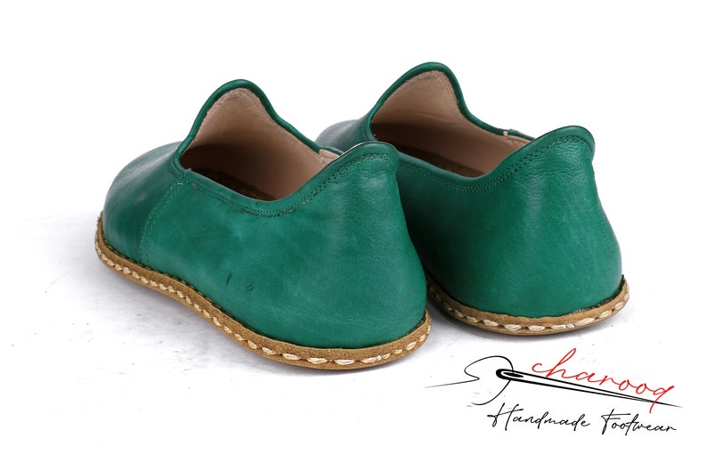 May include: A pair of green leather slip-on shoes with a brown stitched trim. The shoes are handmade and have a casual style.