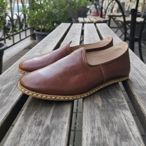 May include: A pair of brown leather slip-on shoes with a rounded toe and a stitched sole. The shoes are made of smooth, polished leather and are displayed on a wooden bench. The shoes appear to be new and unworn.