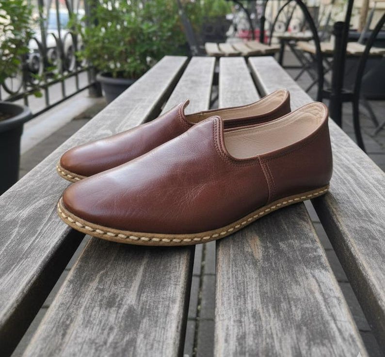 May include: A pair of brown leather slip-on shoes with a rounded toe and a stitched sole. The shoes are made of smooth, polished leather and are displayed on a wooden bench. The shoes appear to be new and unworn.