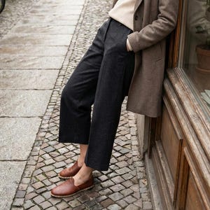 May include: A beige ribbed sweater, a brown wool-blend coat, dark charcoal gray cropped trousers, and brown leather slip-on shoes with a visible stitched jute-style trim along the sole, worn together as a complete outfit ensemble.