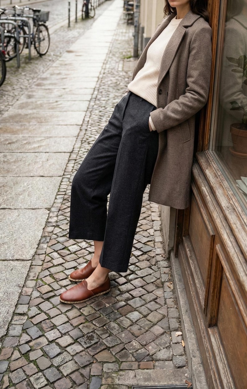 May include: A beige ribbed sweater, a brown wool-blend coat, dark charcoal gray cropped trousers, and brown leather slip-on shoes with a visible stitched jute-style trim along the sole, worn together as a complete outfit ensemble.