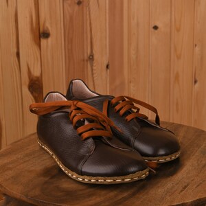 May include: A pair of brown leather lace-up shoes with orange laces. The shoes are sitting on a wooden surface.