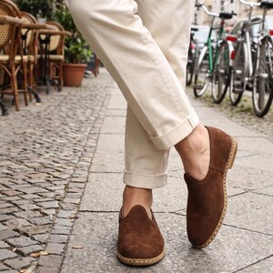 May include: A pair of dark brown suede slip-on shoes with a rounded toe and visible light-colored stitching along the sole, worn with cream-colored cuffed trousers. The shoes feature a low-cut topline and a flat, stacked heel design.