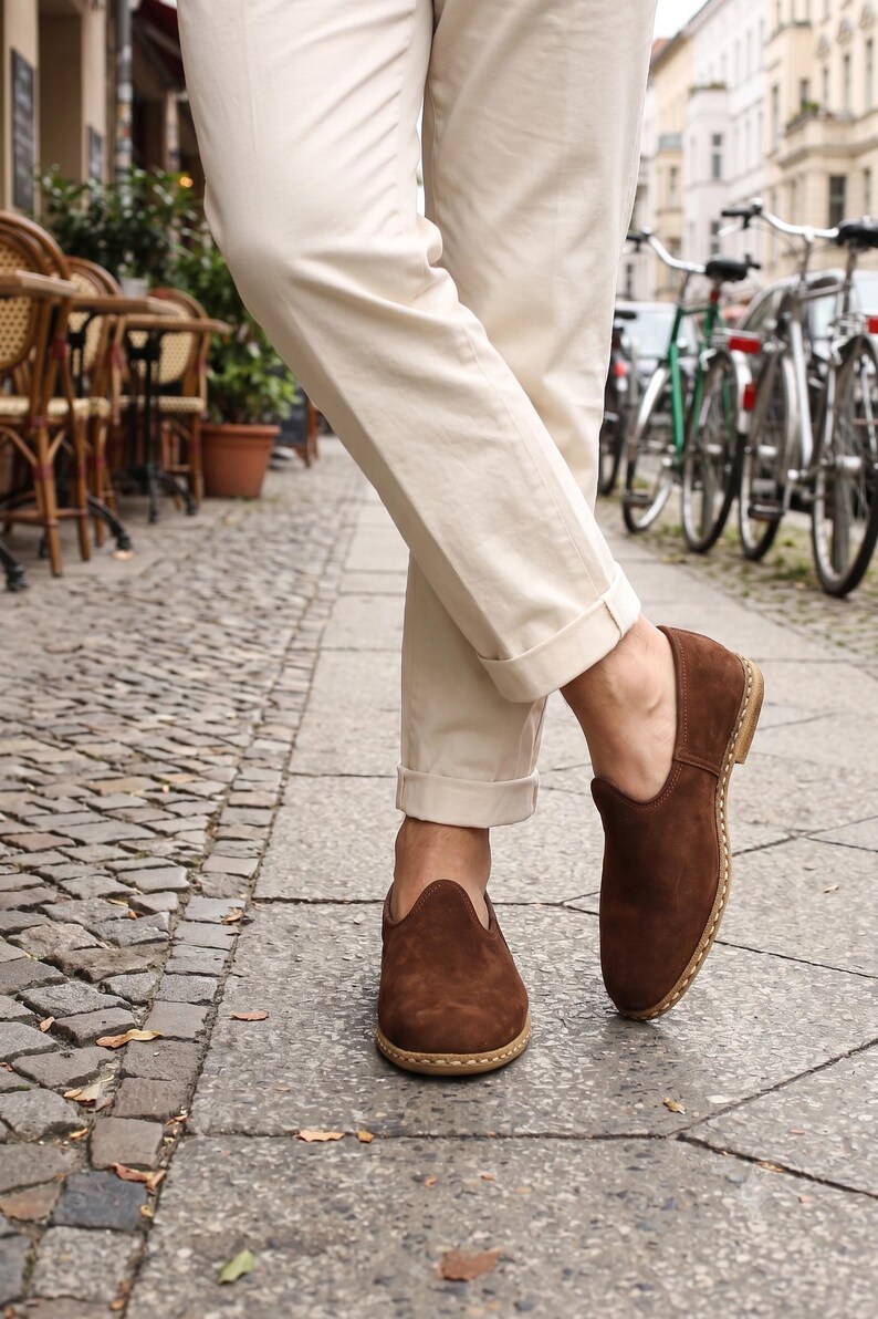 May include: A pair of dark brown suede slip-on shoes with a rounded toe and visible light-colored stitching along the sole, worn with cream-colored cuffed trousers. The shoes feature a low-cut topline and a flat, stacked heel design.