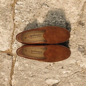 K&ouml;nnte beinhalten: Ein Paar braune Wildleder-Slipper. Die Schuhe haben eine runde Spitze und ein niedriges Profil. Die Innenseite jedes Schuhs hat ein gedrucktes Logo. Die Schuhe werden auf einer Steinoberfl&auml;che pr&auml;sentiert.