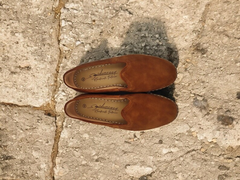 K&ouml;nnte beinhalten: Ein Paar braune Wildleder-Slipper. Die Schuhe haben eine runde Spitze und ein niedriges Profil. Die Innenseite jedes Schuhs hat ein gedrucktes Logo. Die Schuhe werden auf einer Steinoberfl&auml;che pr&auml;sentiert.