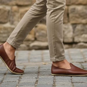 May include: Brown leather slip-on shoes with a stitched sole. The shoes are being worn with khaki pants. The image is taken from a low angle, showing the shoes in motion on a cobblestone surface.
