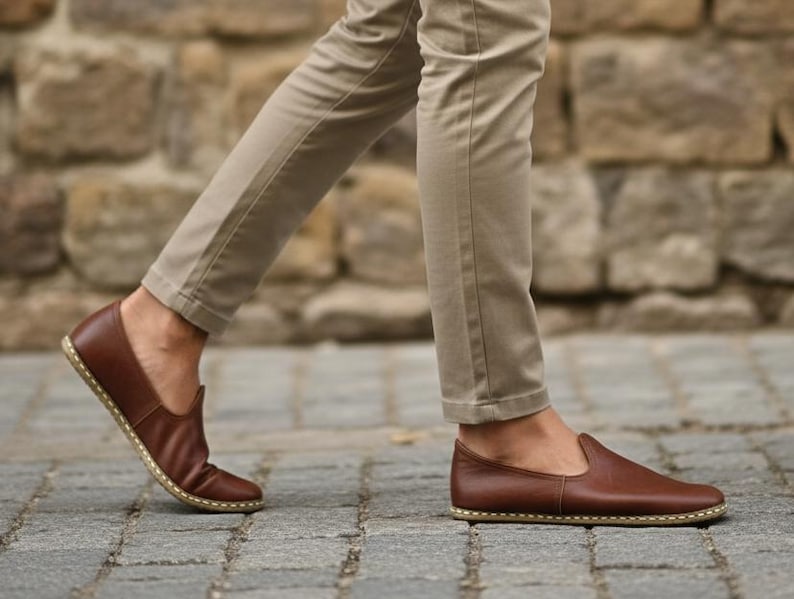 May include: Brown leather slip-on shoes with a stitched sole. The shoes are being worn with khaki pants. The image is taken from a low angle, showing the shoes in motion on a cobblestone surface.