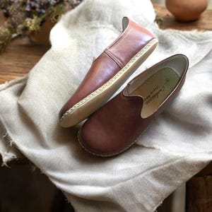 May include: A pair of brown leather slip-on shoes with a stitched edge and a light brown sole. The shoes are on a light brown fabric surface.