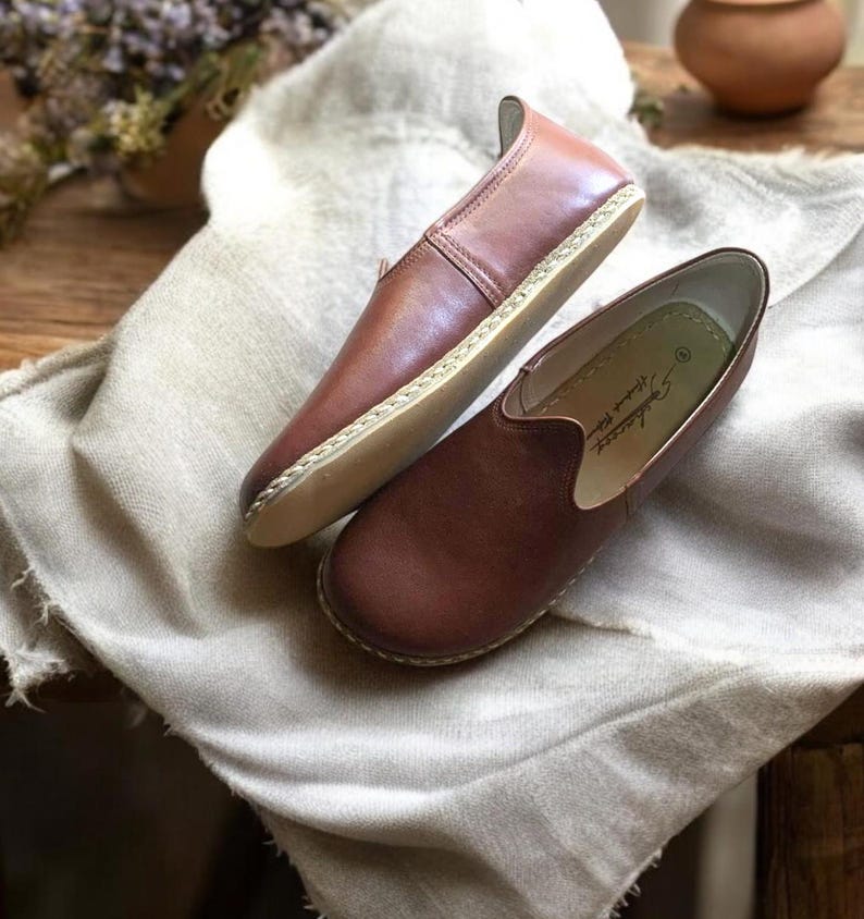 May include: A pair of brown leather slip-on shoes with a stitched edge and a light brown sole. The shoes are on a light brown fabric surface.