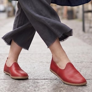 Puede incluir: Zapatos rojos de cuero sin cordones con punta redondeada y suela cosida. Los zapatos son de color rojo vibrante y parecen estar hechos de cuero suave. Los zapatos se muestran en una persona caminando sobre una superficie de hormig&oacute;n.