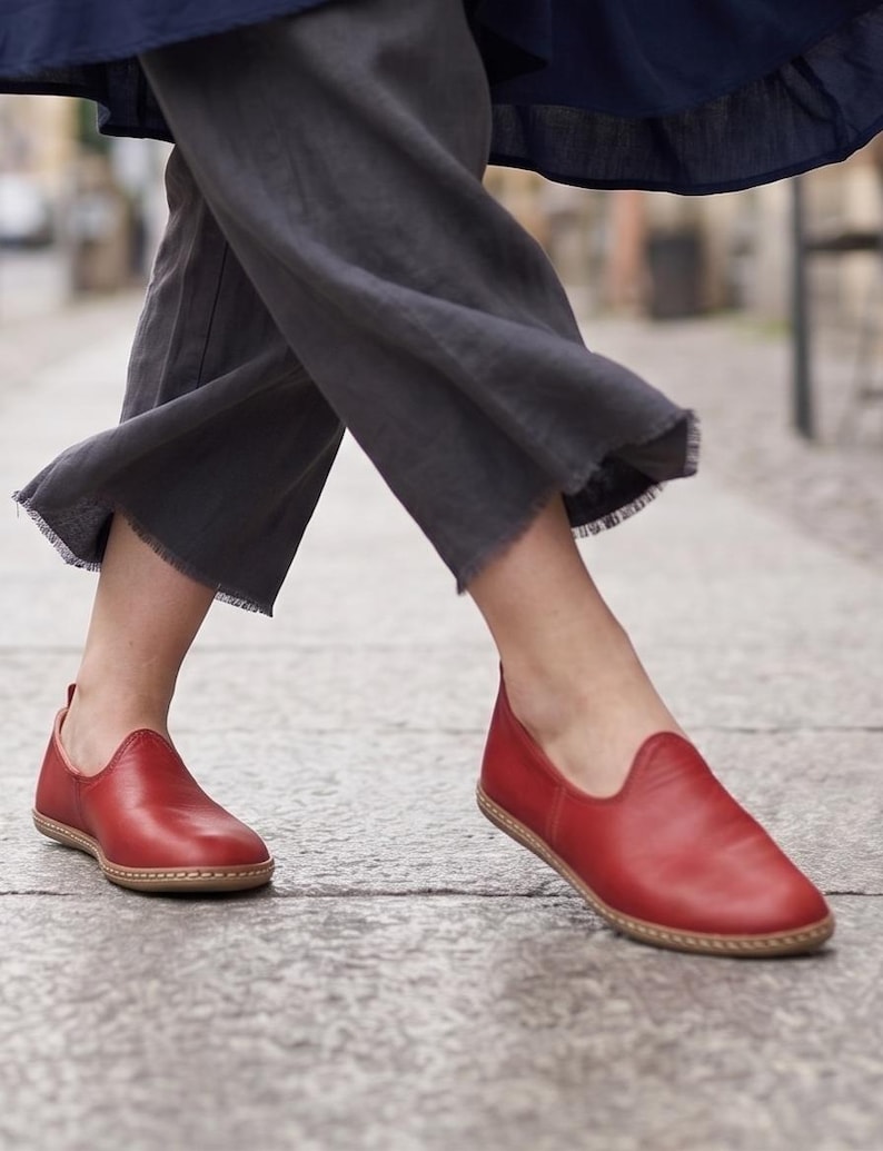 Puede incluir: Zapatos rojos de cuero sin cordones con punta redondeada y suela cosida. Los zapatos son de color rojo vibrante y parecen estar hechos de cuero suave. Los zapatos se muestran en una persona caminando sobre una superficie de hormig&oacute;n.