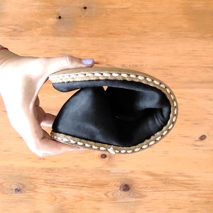 May include: A flexible black shoe with a tan sole and thick white stitching along the edge. The shoe is folded in half to demonstrate its pliability, revealing a smooth black interior lining and a durable, textured outer sole construction.