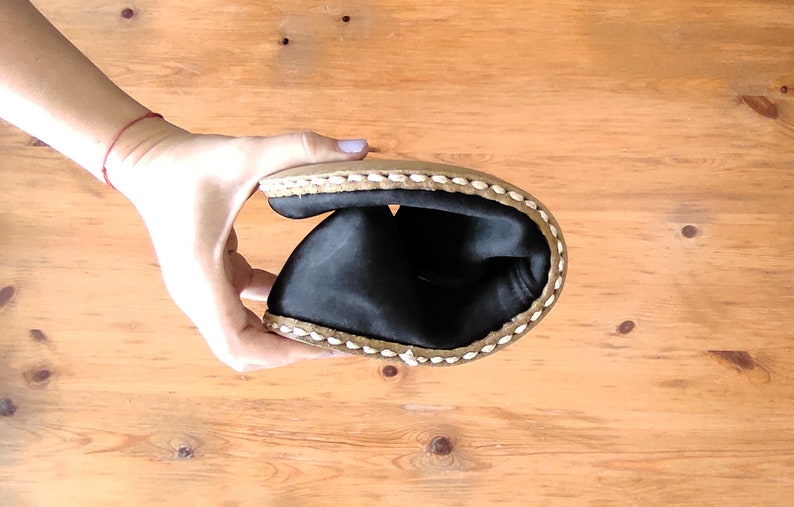 May include: A flexible black shoe with a tan sole and thick white stitching along the edge. The shoe is folded in half to demonstrate its pliability, revealing a smooth black interior lining and a durable, textured outer sole construction.