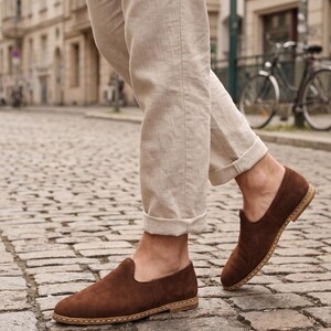 May include: A pair of dark brown suede slip-on shoes featuring a rounded toe, a low-profile silhouette, and a light-colored stitched welt detail along the sole. The shoes are worn with light beige linen trousers that are cuffed at the ankles.