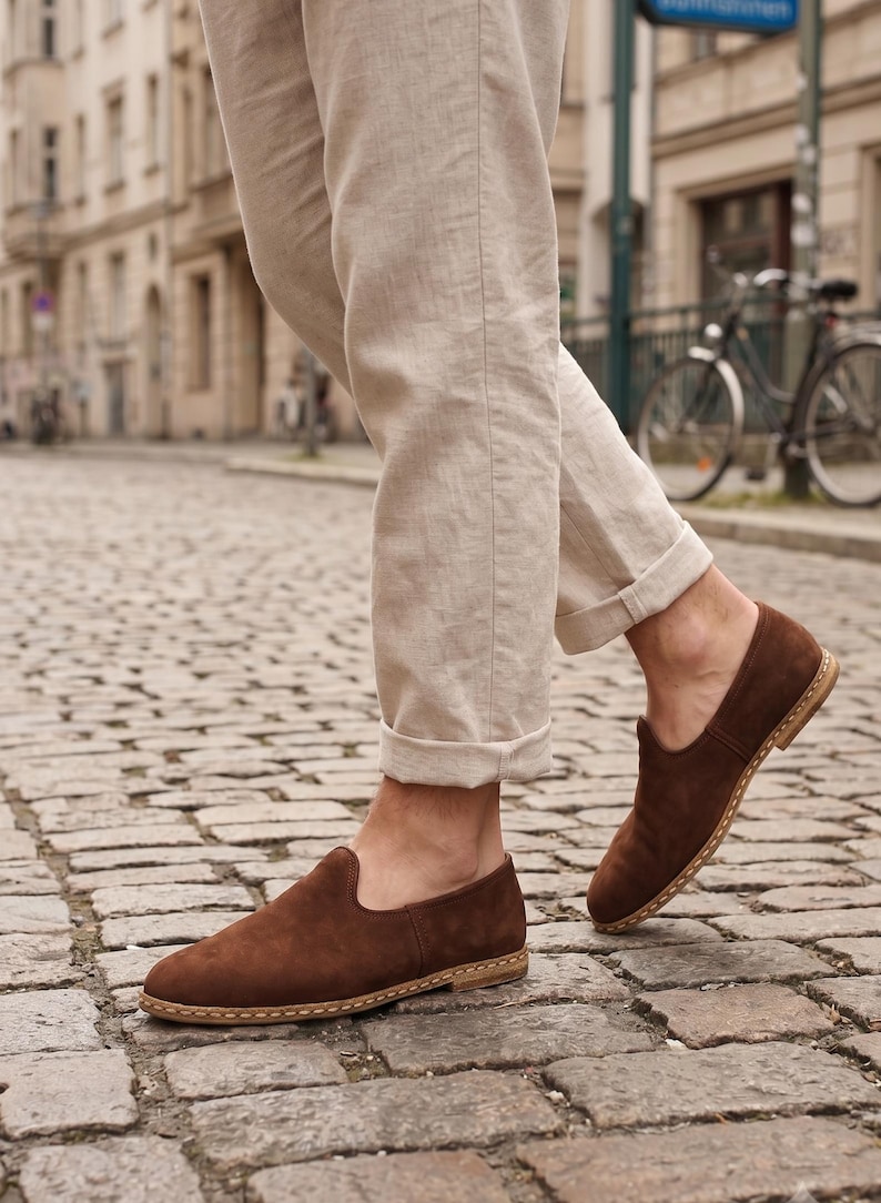 May include: A pair of dark brown suede slip-on shoes featuring a rounded toe, a low-profile silhouette, and a light-colored stitched welt detail along the sole. The shoes are worn with light beige linen trousers that are cuffed at the ankles.