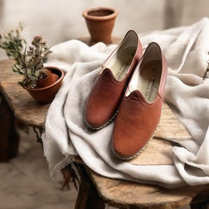 May include: A pair of brown leather slip-on shoes with a rounded toe and stitched sole. The shoes are displayed on a wooden surface with a white cloth. A small terracotta pot with a plant is in the background.
