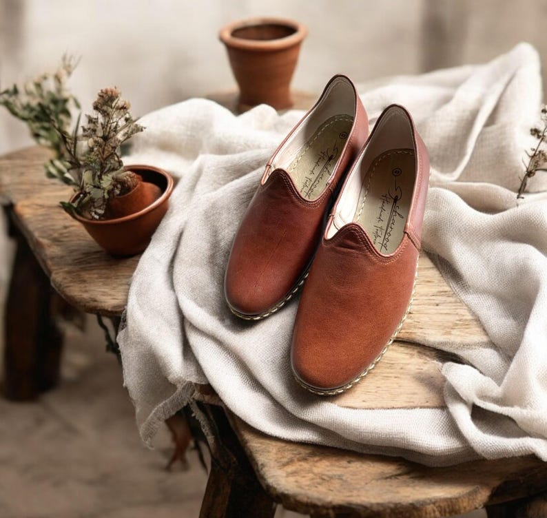 May include: A pair of brown leather slip-on shoes with a rounded toe and stitched sole. The shoes are displayed on a wooden surface with a white cloth. A small terracotta pot with a plant is in the background.