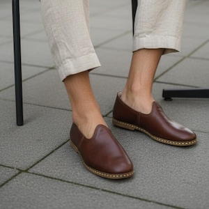 May include: A pair of brown leather slip-on shoes with a rounded toe and a light-colored sole. The shoes are worn by a person wearing beige pants. The shoes appear to be handmade.