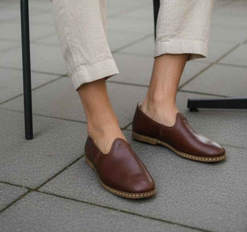 May include: A pair of brown leather slip-on shoes with a rounded toe and a light-colored sole. The shoes are worn by a person wearing beige pants. The shoes appear to be handmade.