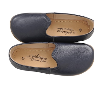 May include: A pair of navy blue slip-on shoes with a textured leather exterior and a tan interior lining. The insole features the text "Scharoog Handmade Footwear" and the number 37. The shoes have a rounded toe and visible stitching along the edge.