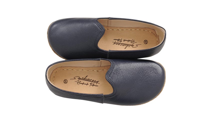 May include: A pair of navy blue slip-on shoes with a textured leather exterior and a tan interior lining. The insole features the text "Scharoog Handmade Footwear" and the number 37. The shoes have a rounded toe and visible stitching along the edge.