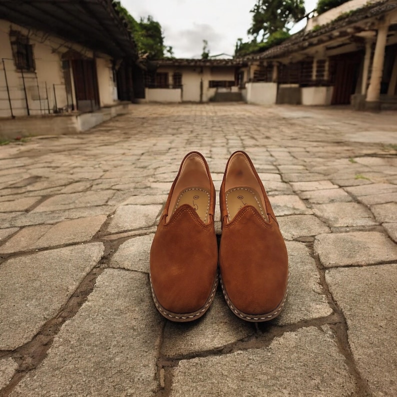 K&ouml;nnte beinhalten: Ein Paar braune Wildleder-Slipper. Die Schuhe haben eine runde Spitze und eine gen&auml;hte Sohle. Der Hintergrund zeigt einen Kopfsteinpflasterweg und Geb&auml;ude.