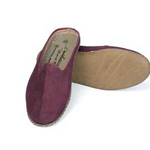 May include: A pair of burgundy suede slippers with a woven jute sole. The slippers have a simple design and are perfect for casual wear.
