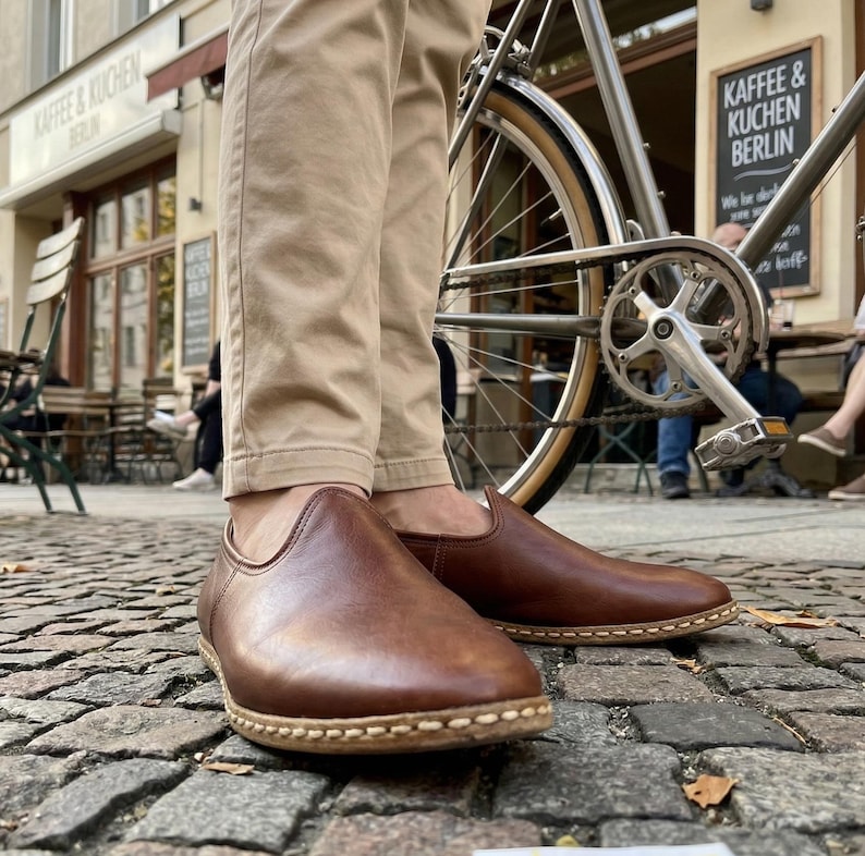 May include: Brown leather slip-on shoes with a rounded toe and stitched sole. The shoes are worn with khaki pants. A bicycle is partially visible in the background, along with a cafe.