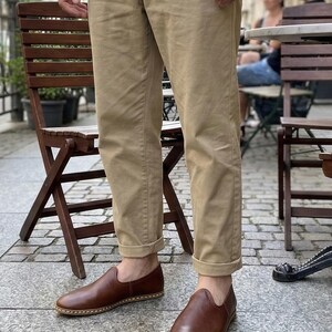 May include: Brown leather slip-on shoes with a rounded toe and a stitched sole. The shoes are worn with khaki-colored pants. The pants are cuffed at the ankles. The person is standing outside on a cobblestone street.