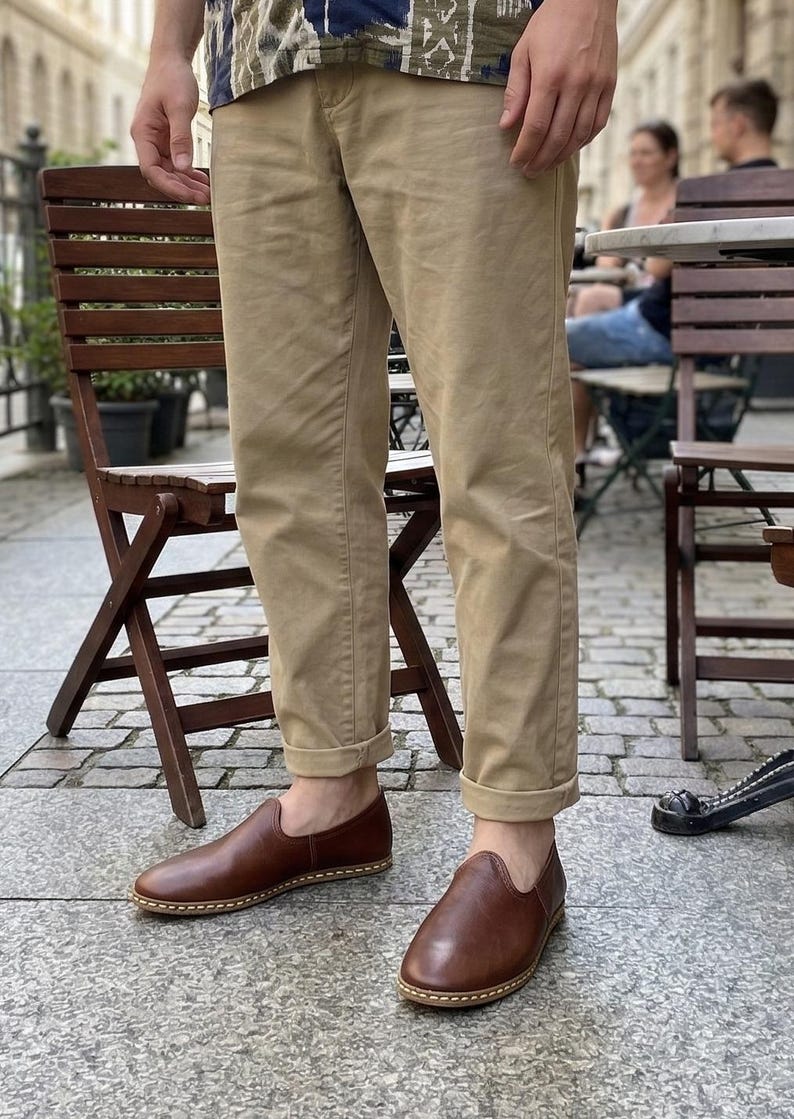 May include: Brown leather slip-on shoes with a rounded toe and a stitched sole. The shoes are worn with khaki-colored pants. The pants are cuffed at the ankles. The person is standing outside on a cobblestone street.