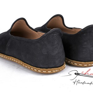 Puede incluir: Un par de zapatos negros de cuero sin cordones con suela cosida marr&oacute;n. Los zapatos son hechos a mano y tienen un aspecto casual y c&oacute;modo.