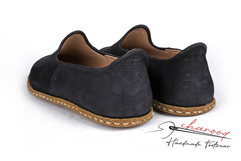 Puede incluir: Un par de zapatos negros de cuero sin cordones con suela cosida marr&oacute;n. Los zapatos son hechos a mano y tienen un aspecto casual y c&oacute;modo.