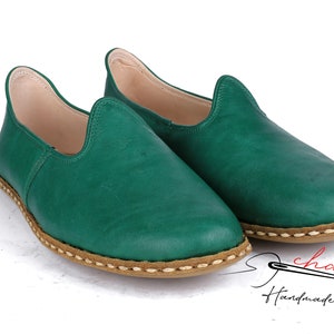 May include: A pair of green leather slip-on shoes with a brown stitched trim. The shoes are handmade and have a casual, comfortable look.