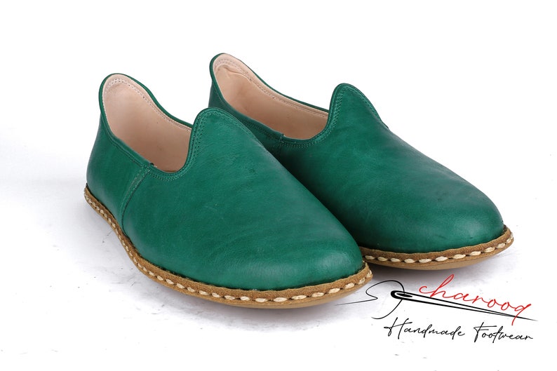 May include: A pair of green leather slip-on shoes with a brown stitched trim. The shoes are handmade and have a casual, comfortable look.