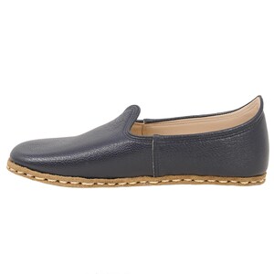 May include: A navy blue slip-on shoe with a textured leather upper and a light tan sole. The shoe features a rounded toe, a low-cut collar, and visible light-colored stitching along the perimeter where the upper meets the flat, flexible sole.