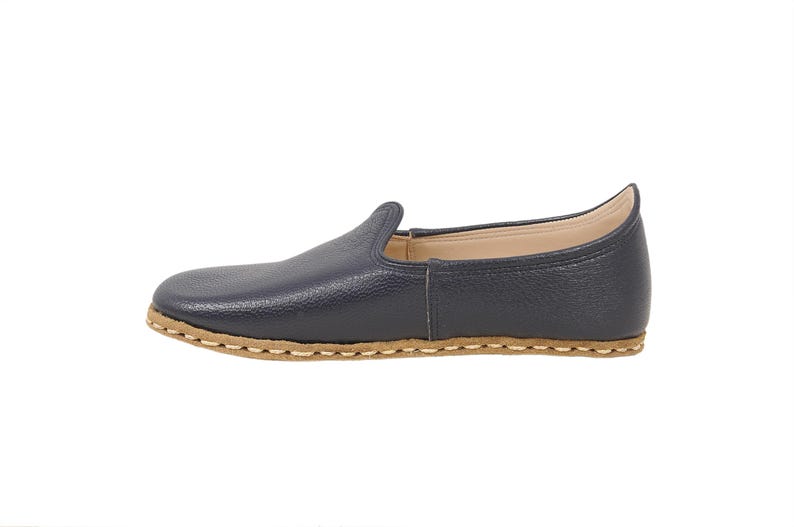 May include: A navy blue slip-on shoe with a textured leather upper and a light tan sole. The shoe features a rounded toe, a low-cut collar, and visible light-colored stitching along the perimeter where the upper meets the flat, flexible sole.