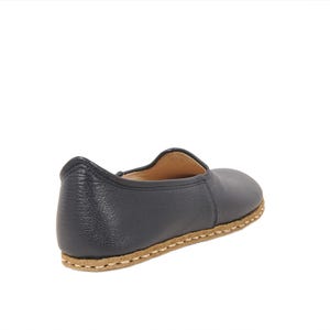 May include: A dark navy blue slip-on shoe with a textured leather upper and a tan-colored sole featuring visible, thick, light-colored stitching around the perimeter. The shoe has a rounded toe, a low-cut collar, and a simple, unadorned design.