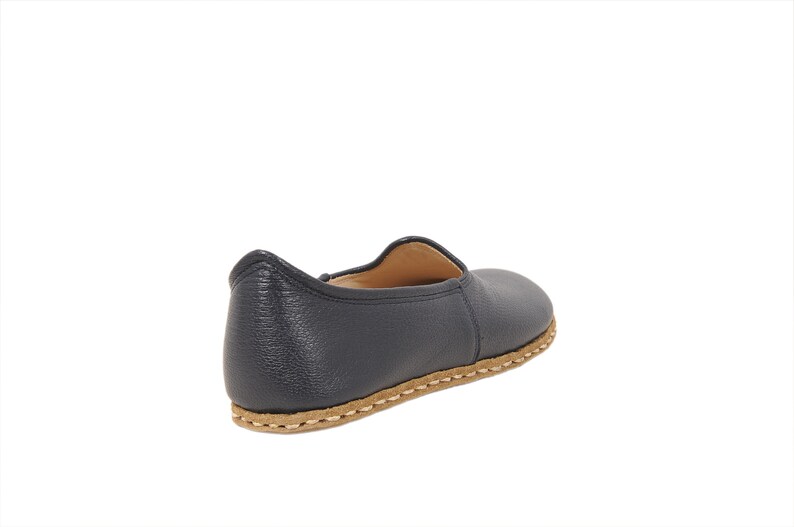 May include: A dark navy blue slip-on shoe with a textured leather upper and a tan-colored sole featuring visible, thick, light-colored stitching around the perimeter. The shoe has a rounded toe, a low-cut collar, and a simple, unadorned design.