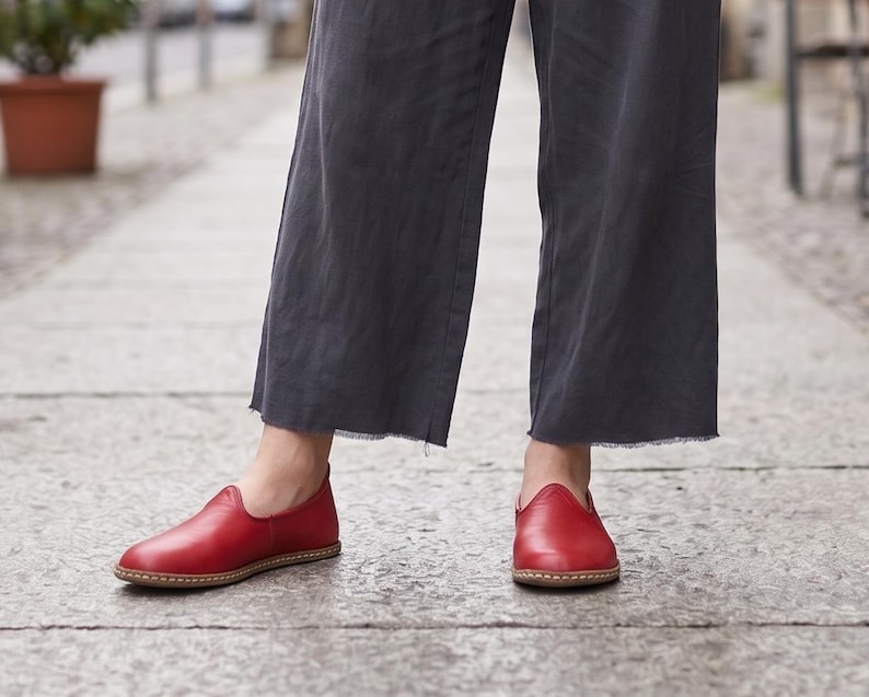 Puede incluir: Un par de zapatos sin cordones de cuero rojo con punta redondeada y suela marr&oacute;n claro. Los zapatos se usan con pantalones anchos grises oscuros. Los zapatos parecen hechos a mano.