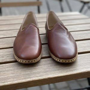 May include: A pair of brown leather slip-on shoes with a rounded toe and stitched sole. The shoes are displayed on a wooden table, showcasing their simple design and neutral color.