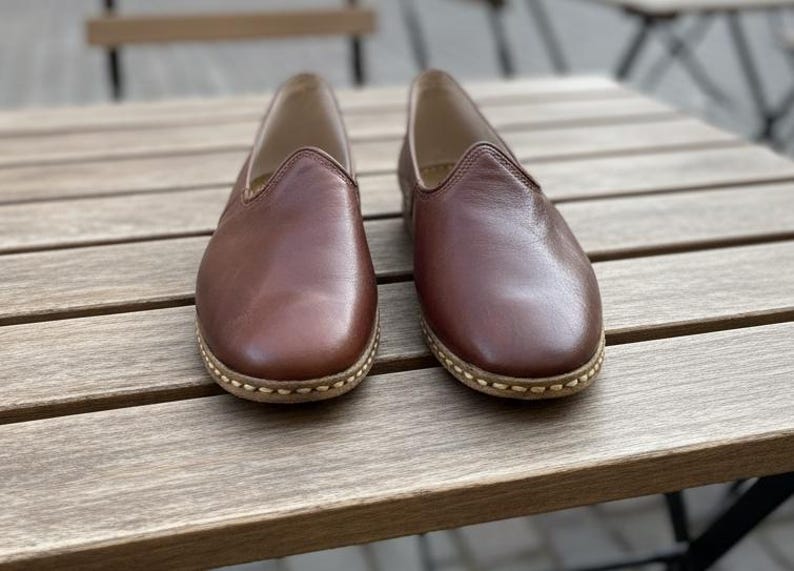 May include: A pair of brown leather slip-on shoes with a rounded toe and stitched sole. The shoes are displayed on a wooden table, showcasing their simple design and neutral color.