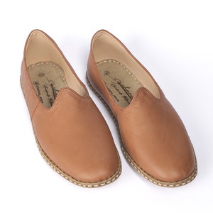 May include: A pair of brown leather slip-on shoes with a stitched edge and a white stitched trim around the sole. The shoes have a label on the inside that reads "Handmade in Morocco".