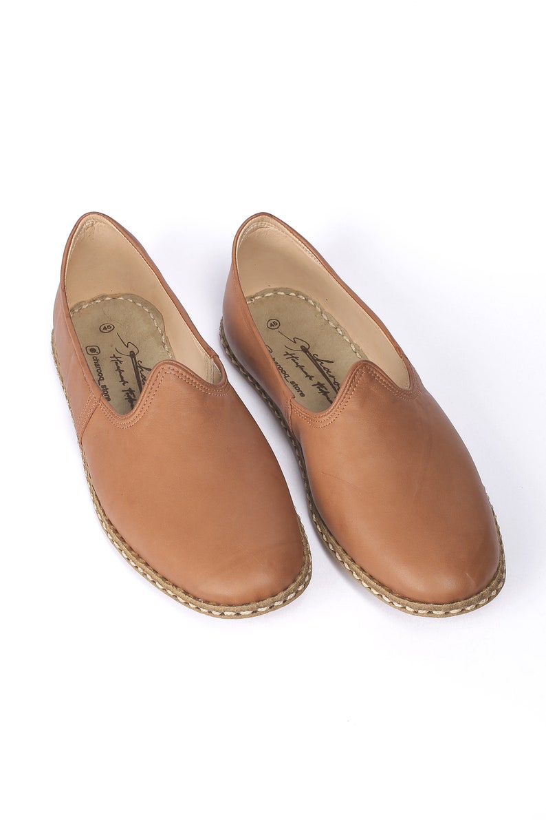 May include: A pair of brown leather slip-on shoes with a stitched edge and a white stitched trim around the sole. The shoes have a label on the inside that reads "Handmade in Morocco".