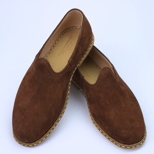 May include: A pair of brown suede slip-on shoes featuring a rounded toe, a tan interior lining, and visible light-colored stitching along the sole edge. The interior sole is stamped with the text TARIK KOSGER 0536 445 03 38 KU 13.