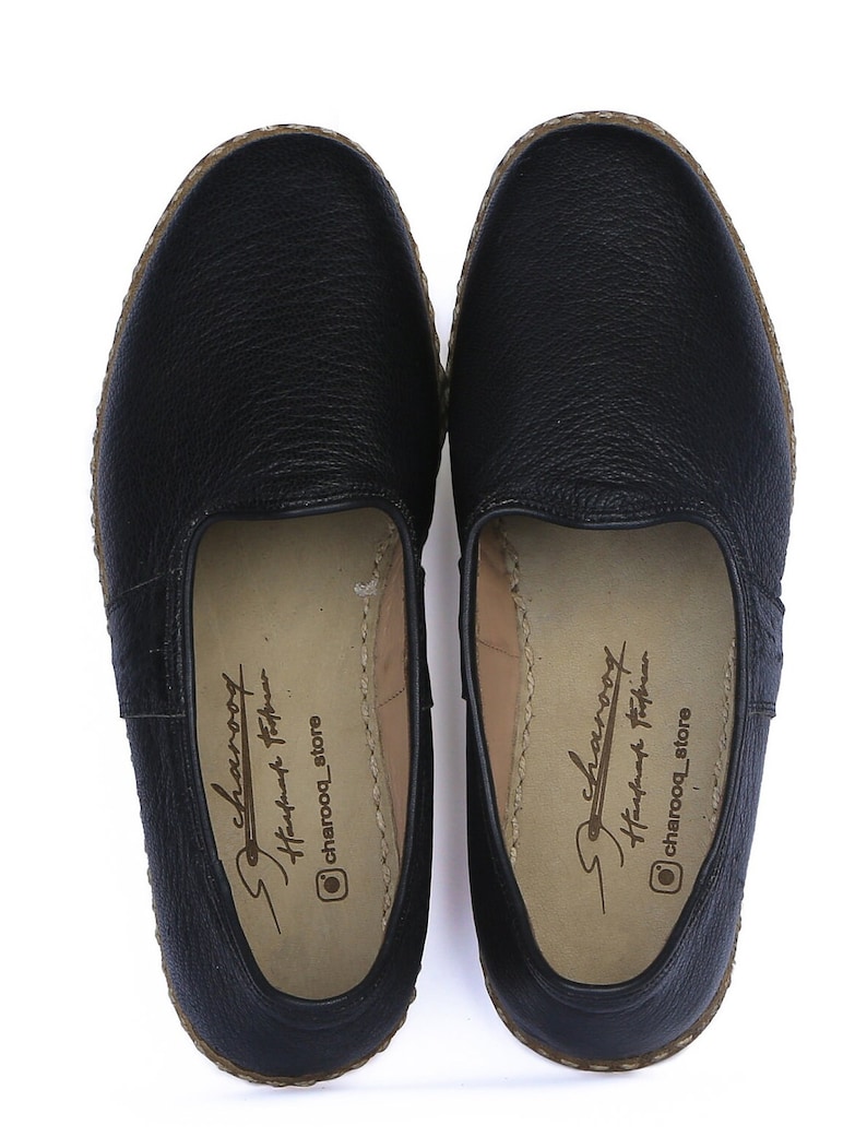 Op de afbeelding: Een paar zwarte leren instapschoenen met een bruine geweven rand. De schoenen hebben een "Charooq Handmade Footwear" label aan de binnenkant.