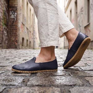 May include: A pair of dark navy blue slip-on shoes with a textured leather upper and a tan-colored sole featuring visible light-colored stitching along the perimeter. The shoes have a rounded toe and a low-cut profile, showing a simple construction.
