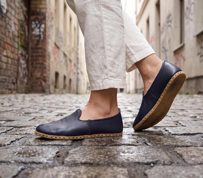 May include: A pair of dark navy blue slip-on shoes with a textured leather upper and a tan-colored sole featuring visible light-colored stitching along the perimeter. The shoes have a rounded toe and a low-cut profile, showing a simple construction.