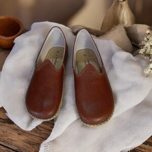May include: A pair of brown leather slip-on shoes with a pointed toe and a stitched edge. The shoes are on a white fabric surface.