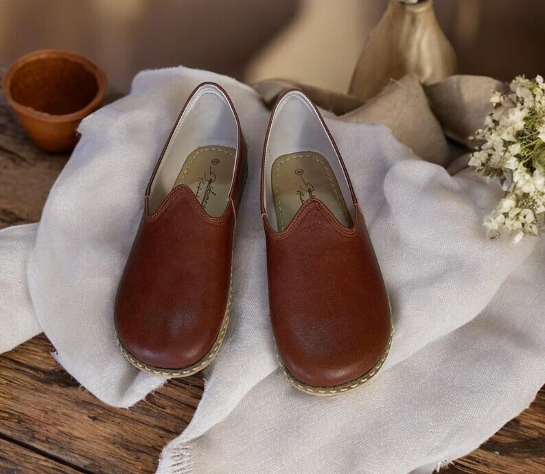 May include: A pair of brown leather slip-on shoes with a pointed toe and a stitched edge. The shoes are on a white fabric surface.